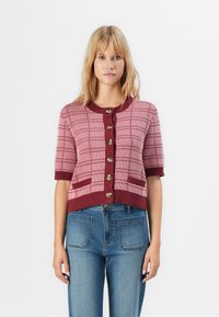 Femme blonde portant un cardigan court à manches courtes à carreaux roses et bordeaux avec de grands boutons, et un jean bleu avec des poches avant.
