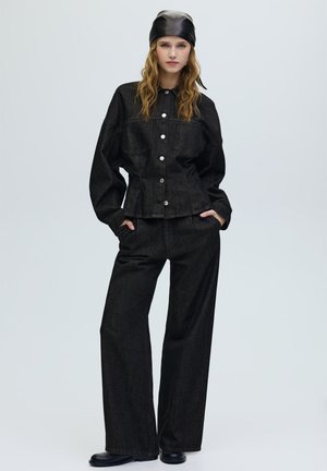 Sort denimjakke med sølvknapper og to brystlommer, kombineret med sorte bukser med vide ben. Modellen bærer et sort læderhårbånd.