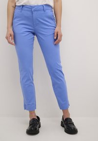 Helleblaue, knöchellange Hosen mit einer glatten Textur, die einen Frontreißverschluss und einen Knopfverschluss sowie Seitentaschen aufweisen. Tragen mit schwarzen Schuhen.