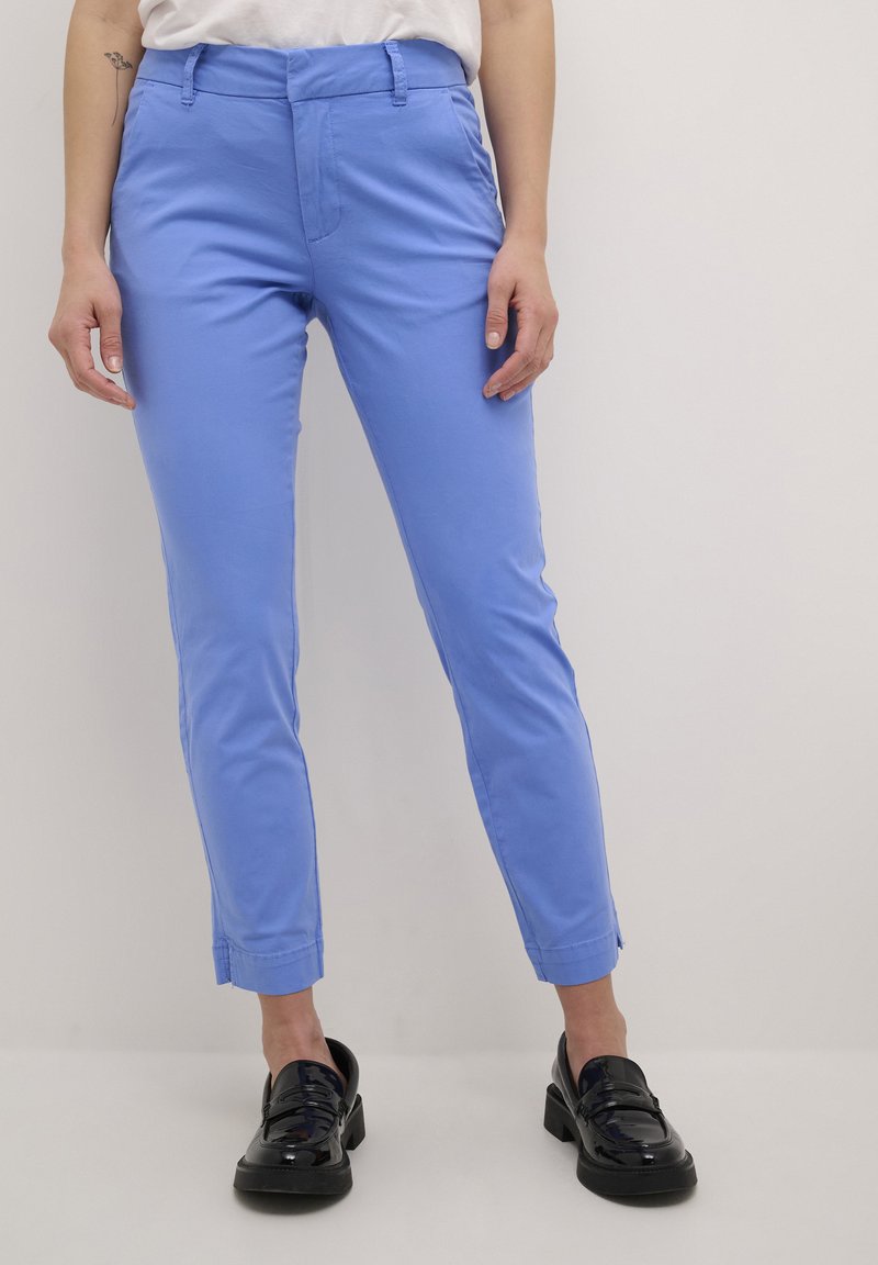 Pantalons blue clair à la cheville, avec une texture lisse, dotés d'une fermeture zippée et à bouton à l'avant, ainsi que de poches latérales. Portés avec des chaussures noires.