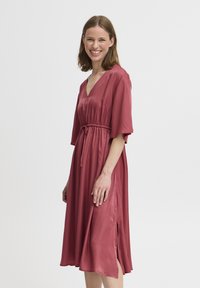 Robe rose avec un décolleté en V, des manches courtes, une taille à nouer et des fentes sur les côtés. Fabriquée dans un tissu lisse et fluide avec une coupe décontractée.