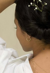 Pendientes de aro de oro con un diseño retorcido, llevados por una persona con cabello oscuro, adornada con pequeñas flores blancas y una camisa blanca.