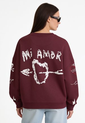 Femme avec des lunettes de soleil portant un sweat bordeaux avec « Mi Amor » et un cœur transpercé par une flèche imprimés dans le dos, jean pâle, fond uni.