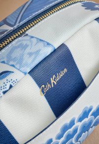 Zila un baltā rakstainā kosmētikas soma ar teksturētu virsmu, zelta rāvējslēdzis un zelta "Cath Kidston" logo, kas izceļ viļņotu dizainu.