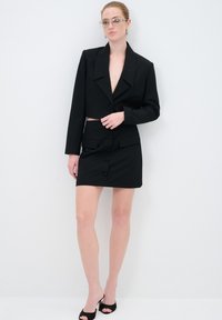 Zwarte cropped blazer met een diepecolleté, gecombineerd met een bijpassende zwarte mini-jurk met knoopdetails en twee voorzakken.