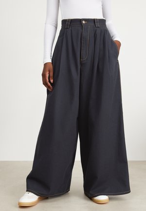 Osoba nosząca czarne spodnie z szerokimi nogawkami i wysokim stanem, z przednimi plisami, białą bluzkę z długim rękawem oraz białe trampki z gumowymi podeszwami.