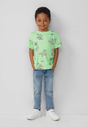 Grünes T-Shirt mit kurzen Ärmeln, auf dem Cartoon-Dinosaurier auf Surfbrettern abgebildet sind; kombiniert mit hellblauen Jeans und weißen Sneakers. Glattes, lässiges Material.