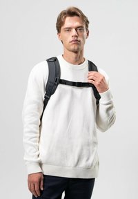 Mochila negra con correas ajustables llevada sobre un suéter blanco de punto acanalado. La figura está de pie contra un fondo neutro.