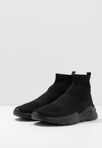 Černé pletené tenisky s designem jako ponožky, texturovaným svrškem, vysokým opatkem a odolnou gumovou podrážkou. Minimalistický styl bez viditelných značek.