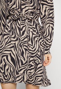 Vestido com estampa de zebra em bege e preto, com cintura amarrada, mangas longas e uma barra com babados, feito de um tecido texturizado.