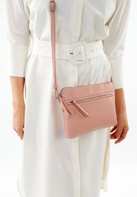 Borsa a tracolla in pelle rosa con superficie texturizzata, dotata di due scomparti con zip e una tracolla removibile, indossata su un vestito bianco.