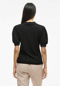 Blusa nera con texture, maniche a sbuffo corte, scollatura rotonda e silhouette aderente, abbinata a pantaloni beige chiaro sartoriali.