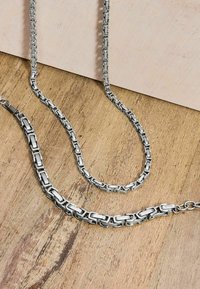 Collane in metallo argentato con catene a maglie rettangolari, di lunghezze variabili, esposte su una superficie di legno. Texture liscia e finitura lucida.