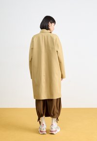Manteau long beige avec col, doté d'une couture de pince au dos, associé à un pantalon ample marron et des chaussures de sport avec des accents pastel.