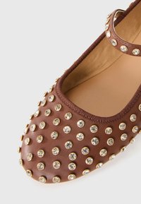 Chaussure plate marron à bout rond ornée de multiples strass transparents fixés sur des clous dorés et d'une bride de cheville assortie.