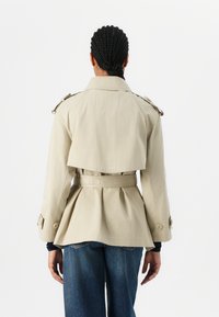 Beige trench coat with a belted waist, back pleats, and shoulder epaulets. Features long sleeves and a pointed collar. Denim jeans visible below.