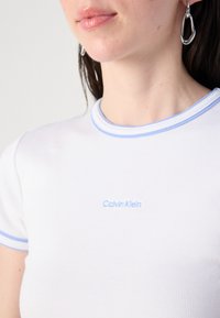Witte geribde t-shirt met lichtblauwe afwerking en geborduurd "Calvin Klein" logo op de borst. Soepele textuur, aansluitend ontwerp.