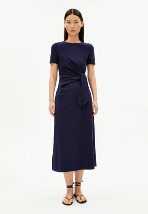 Vrouw draagt een marineblauwe midi-jurk met korte mouwen en een knoopdetail in de taille, gecombineerd met zwarte platte sandalen met bandjes, en staat rechtop.