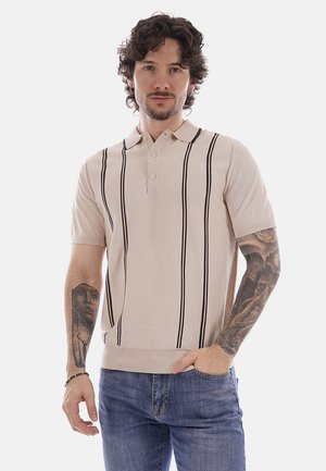 Uomo con capelli ricci e tatuaggi indossa una polo beige a maniche corte con strisce verticali nere e jeans blu, in piedi con una mano in tasca.