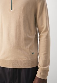 Beżowy sweter dzianinowy z kołnierzem na pół zamku, prążkowanymi mankietami i dołem. Małe zielone logo "PS" w dolnym lewym rogu. Miękka faktura.