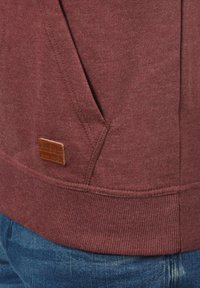 Maroon sweatshirt with textured fabric, ribbed hem and cuffs, featuring a small brown leather logo tab near the pocket.