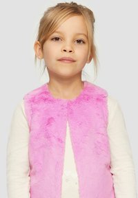 Gilet en fausse fourrure rose à la texture douce, avec un design sans col, porté par-dessus un haut à manches longues blanc avec des accents en forme de cœur.