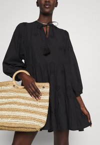 Black dress with puff sleeves, embroidered details, and a tie at the neck. Holding a striped straw bag with a woven texture and leather accents.