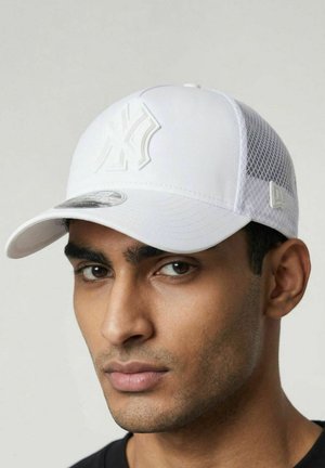 Junger Mann mit mittlerem Hautton, der eine weiße Netz-Baseballmütze mit einem hervorgehobenen Logo auf der Vorderseite trägt und nach vorne schaut.