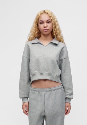 Young woman with long curly blonde hair wearing a gray cropped sweatshirt and matching gray sweatpants against a plain background.