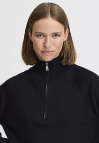 Pullover nero a mezza zip realizzato in morbido materiale a maglia. Presenta un colletto alto, maniche raglan e una texture liscia. Dettaglio della cerniera in argento.