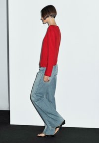 Ein Modell in einem roten Pullover und weit geschnittenen blauen Jeans läuft vor einem schlichten weißen Hintergrund und hebt einen modernen Mode-Look hervor.