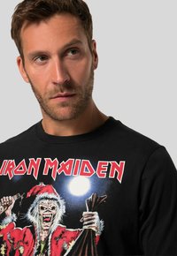 Černá mikina s grafickým potiskem monstra v červeném kabátu s bílým kožešinovým límcem, které drží tašku, a s textem "IRON MAIDEN."