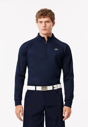 Marineblauwe longsleeve polo met kwart-rits, gewatteerde stof, groene logo op de borst, gecombineerd met donkere broeken en een witte riem met een zilveren gesp.