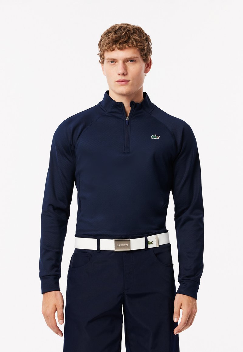 Polo de manga larga azul marino con cremallera de cuarto, tejido texturizado, logo verde en el pecho, combinado con pantalones oscuros y un cinturón blanco con una hebilla plateada.