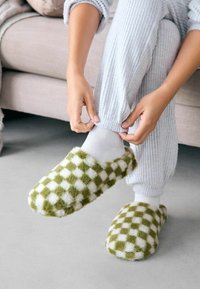Furry green and white checkered slippers made of plush material, worn with light grey, ribbed pants. Soft, rounded design.