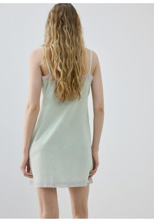 Vrouw met lang golvend haar, gekleed in een lichtgroene mouwloze jurk met kanten rand, staand met het gezicht naar een eenvoudige lichtgrijze achtergrond.