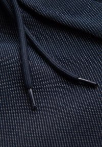 Tessuto navy con una texture e piccoli punti più chiari, caratterizzato da cordini neri con punte in metallo. Il dettaglio in primo piano mette in risalto il motivo e il materiale.