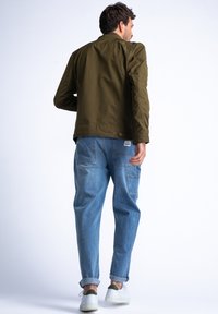 Giubbotto bomber verde oliva con un design aderente, abbinato a pantaloni in denim azzurro chiaro e sneakers bianche. Vista posteriore semplice.
