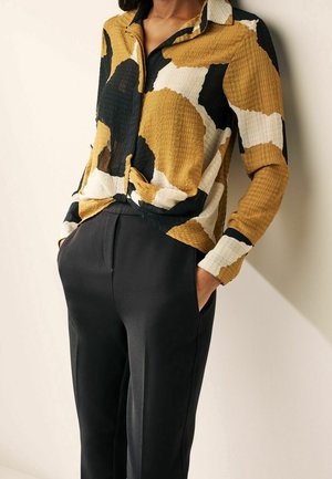 Persona que lleva una camisa de manga larga con un patrón en mostaza, negro y blanco, metida dentro de unos pantalones negros de talle alto con las manos en los bolsillos.