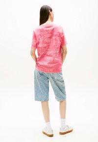 Różowa, mineralnie zafarbowana koszulka typu T-shirt z krótkim rękawem, z białymi logo na plecach. Zestawiona z jasnoniebieskimi szortami dżinsowymi i białymi sneakersami.