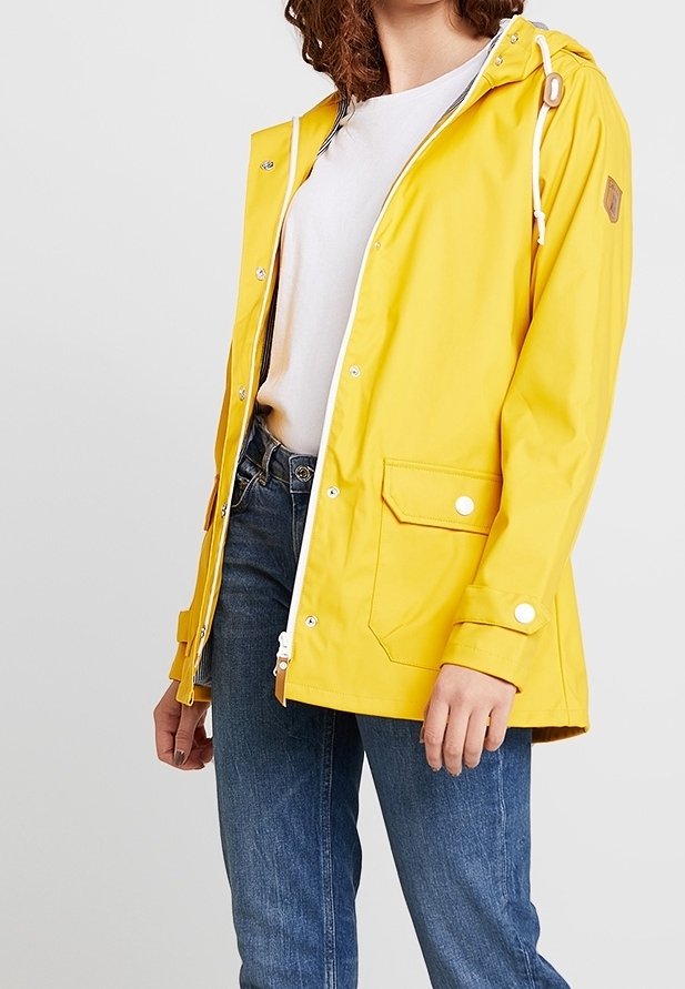 Chaqueta amarilla impermeable con capucha, cremallera blanca y regulador ajustable. Cuenta con bolsillos delanteros y botones a presión. Llevada sobre una camiseta blanca.
