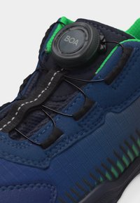 Κοντινό πλάνο ενός μπλε αθλητικού παπουτσιού με πράσινες λεπτομέρειες, που διαθέτει μαύρο σύστημα κορδονιών BOA με δύο λεπτά μαύρα σύρματα για σύσφιξη.