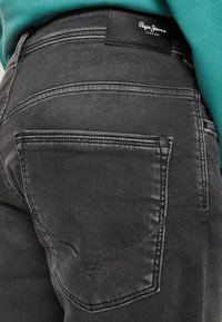 Vaqueros de mezclilla negra con una textura ligeramente desgastada, que cuentan con un bolsillo trasero con costuras en contraste y una etiqueta de marca de cuero negro en la cintura.