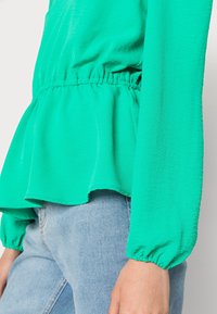 Blusa verde a maniche lunghe con vita arricciata, tessuto fluido e polsini elasticizzati, abbinata a jeans in denim blu chiaro.