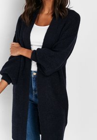 Cardigan tricoté bleu marine avec un design texturé à ouverture frontale et des manches surdimensionnées. Porté sur un haut blanc et associé à un jean en denim.