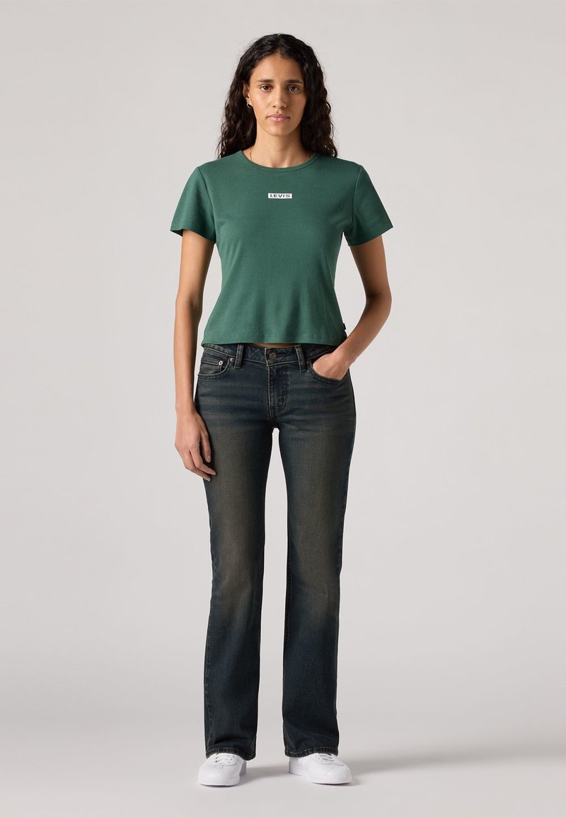 T-shirt vert à coupe courte avec col rond et logo, associé à un jean droit en denim foncé et des baskets blanches. Le modèle se tient avec les mains dans les poches.