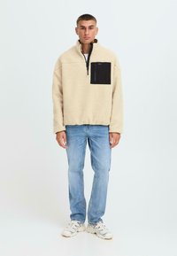 Sudadera de forro polar beige con cuello de media cremallera, bolsillo en el pecho negro y corte casual. Combinada con jeans de color azul claro y zapatillas blancas.