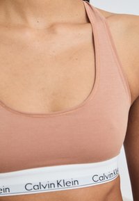 Világos rózsaszín sportmelltartó, puha anyagból készült, lekerekített nyakkivágással, széles fehér pánttal, az alján fekete "Calvin Klein" logóval.