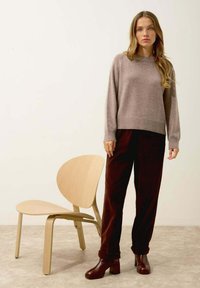 Jasnobrązowy sweter dziergany z prążkowanymi mankietami, połączony z ciemnobrązowymi spodniami z cordurowej tkaniny i masywnymi, brązowymi botkami na obcasie. W tle drewniane krzesło.