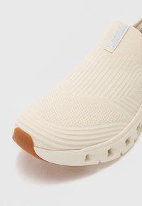 Béžová nazouvací bota s texturovaným pleteným svrškem, ribbovaným designem a hnědou gumovou špičkou; obsahuje výřezy v podrážce pro ventilaci.